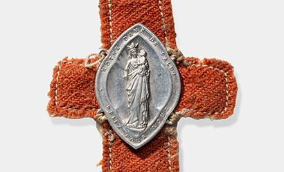 Modern photograph of a silver-colored oval medal depicting a woman in robes holding an infant. The medal has been sewn onto a brown cloth cross.