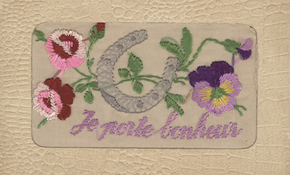 Scan of the front of a postcard with silk embroidery depicting a horseshoe and several flowers. Embroidered text in purple: 'Je porte bonheur'