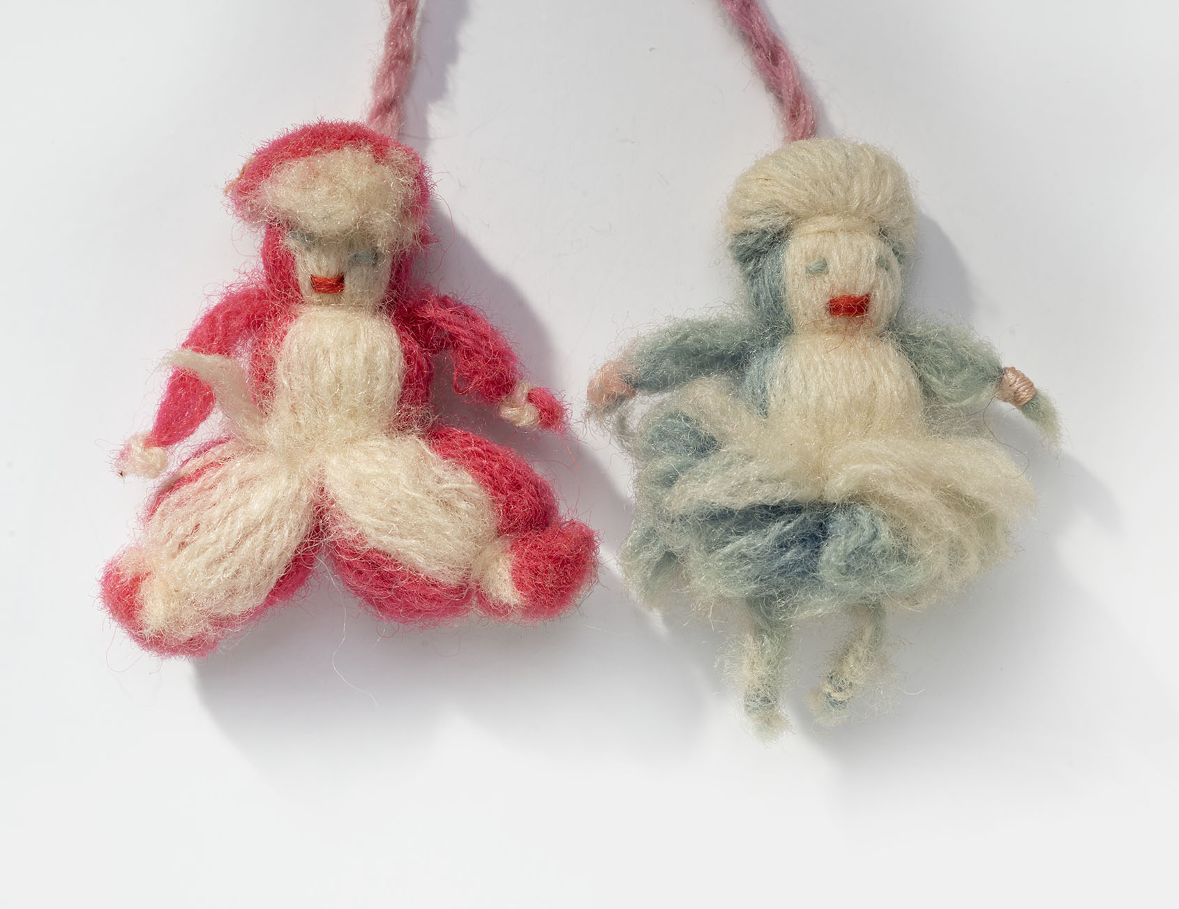 Two doll charms made out of yarn. Left doll charm is red and cream-colored yarn and wears pants. Right doll charm is blue and cream-colored yard and wears a skirt.