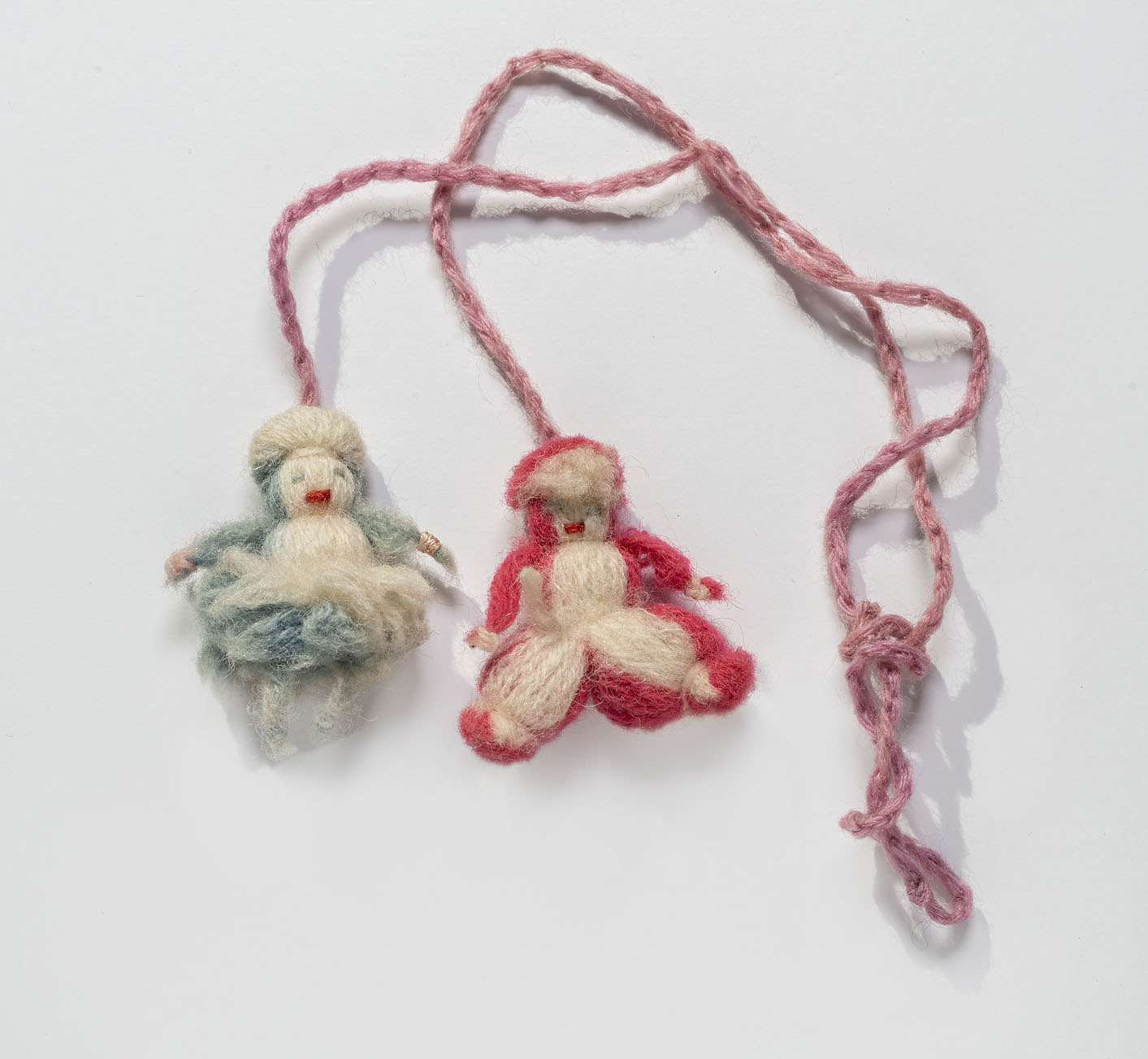 Two doll charms made out of yarn attached to each other with yarn strings. One doll charm is red and cream-colored yarn and wears pants. The other doll charm is blue and cream-colored yard and wears a skirt.