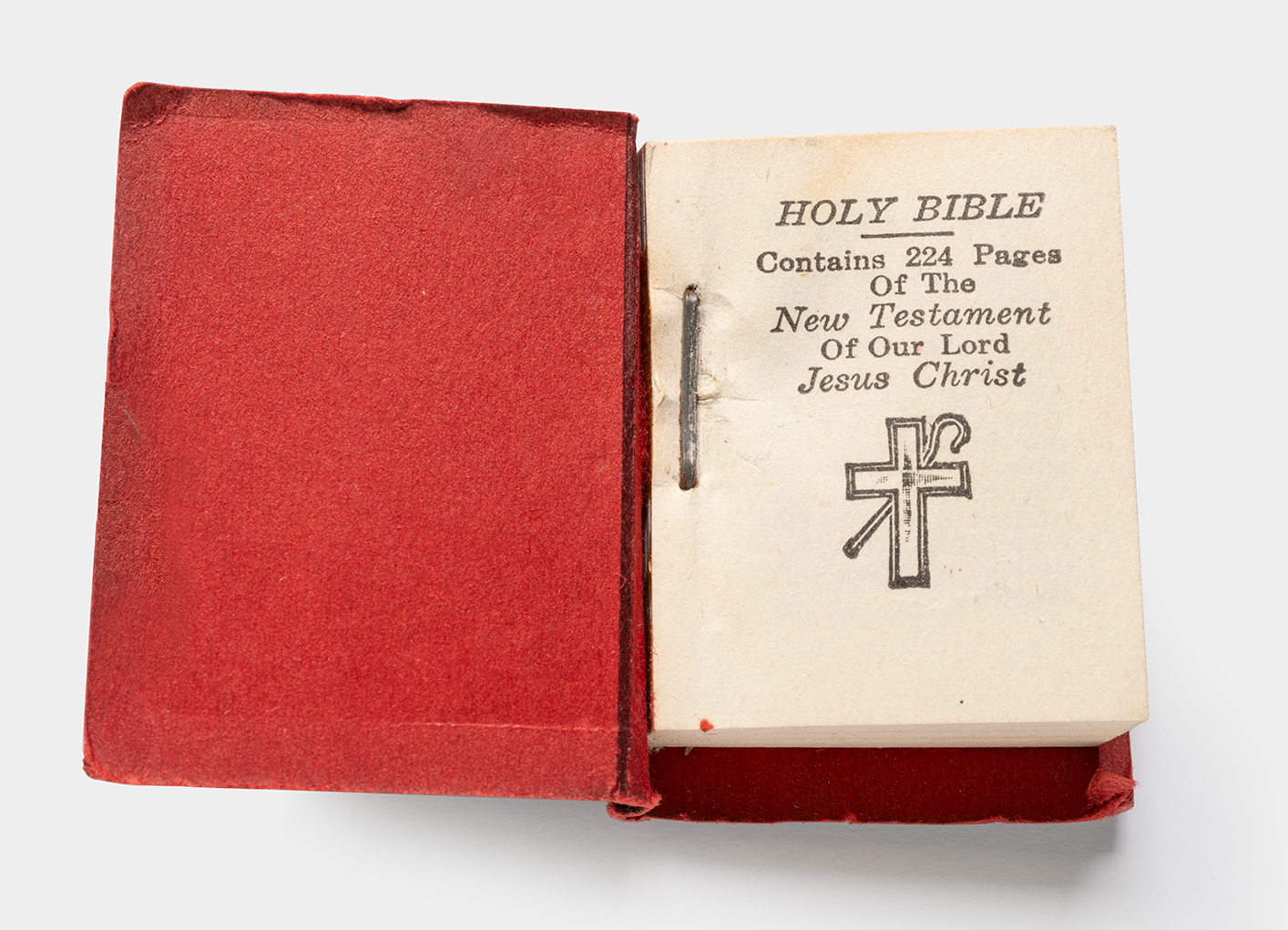 A miniature Bible with the front cover open. The inner lining is red. The front page has typewritten text and an illustration of a cross and shepherd's hook. Text: 'HOLY BIBLE / Contains 224 Pages Of The New Testament Of Our Lord Jesus Christ'