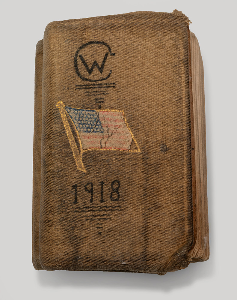 Closed book, battered and worn. Front cover adorned with a stylized handwritten 'CW', a U.S. flag and a handwritten '1918'