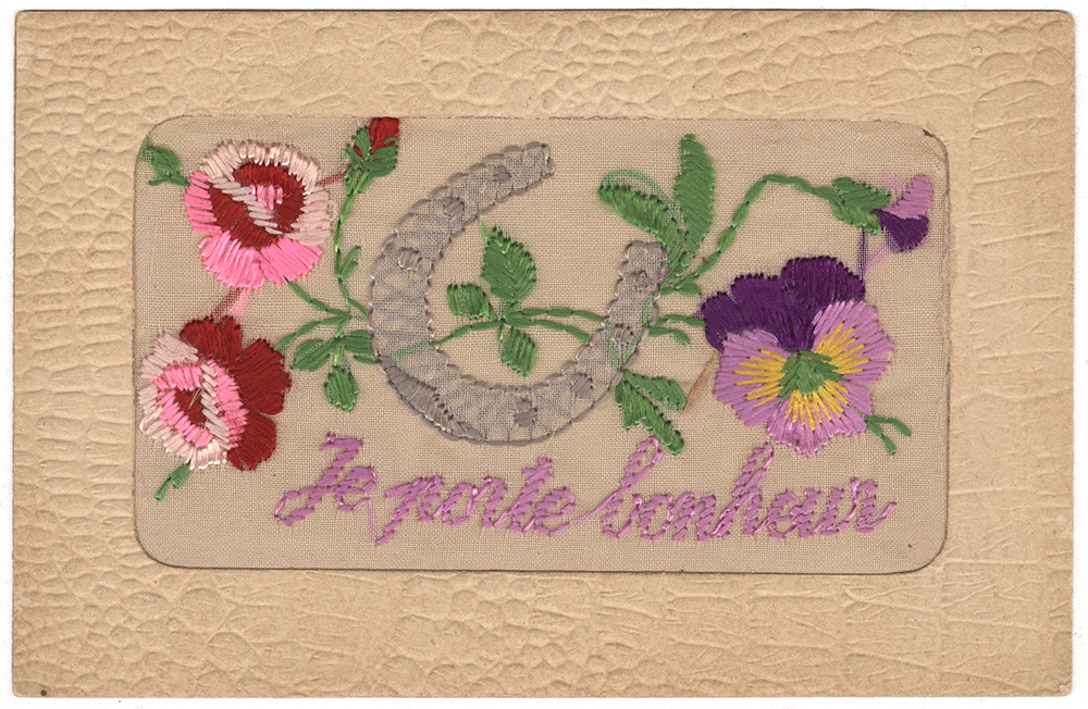 Scan of the front of a postcard with silk embroidery depicting a horseshoe and several flowers. Embroidered text in purple: 'Je porte bonheur'