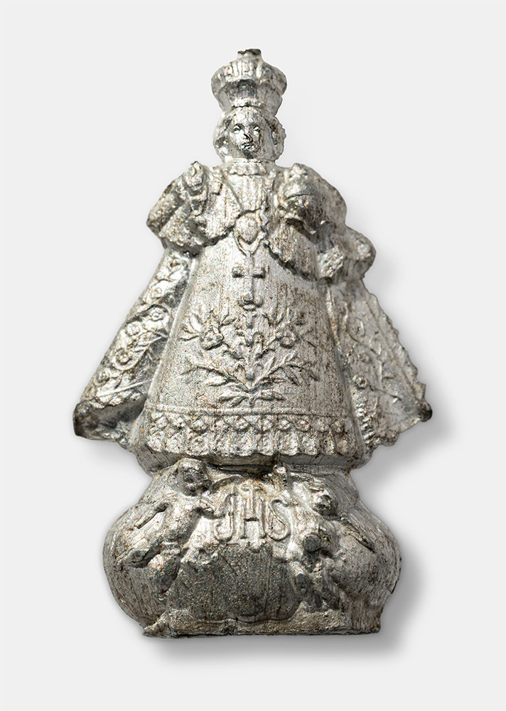 Silver-colored figurine of a boy dressed in elaborate kingly robes and crowned with a hoop crown.