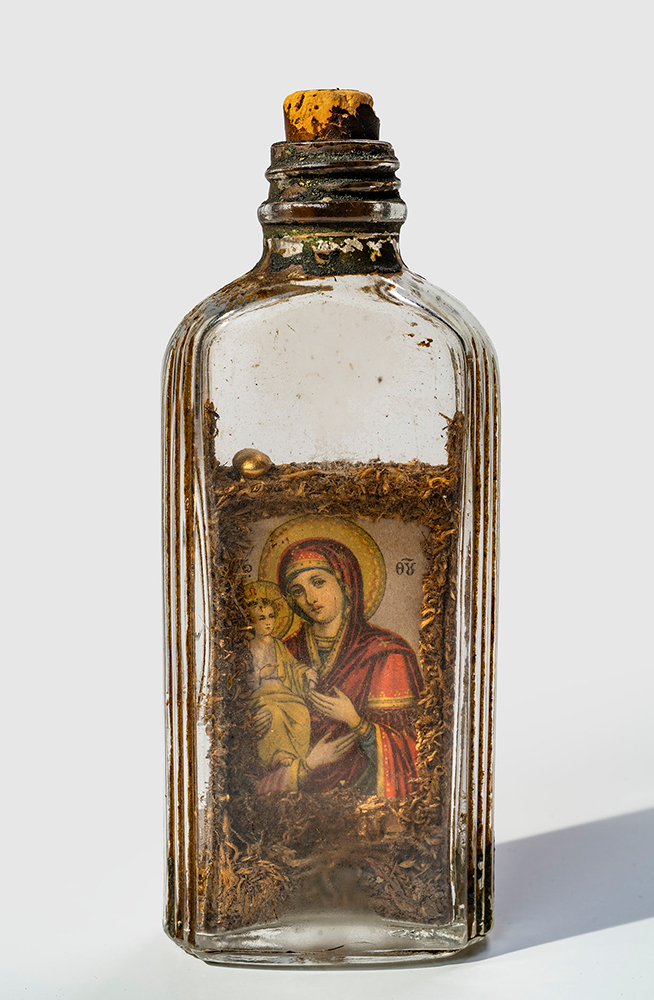 Modern photograph of a glass bottle with cork stopper. One side displays the image of Madonna and Christ Child. Images are decorated with dried tobacco on all sides. A small golden bead sits on top of the icons and it appears others used to be present.