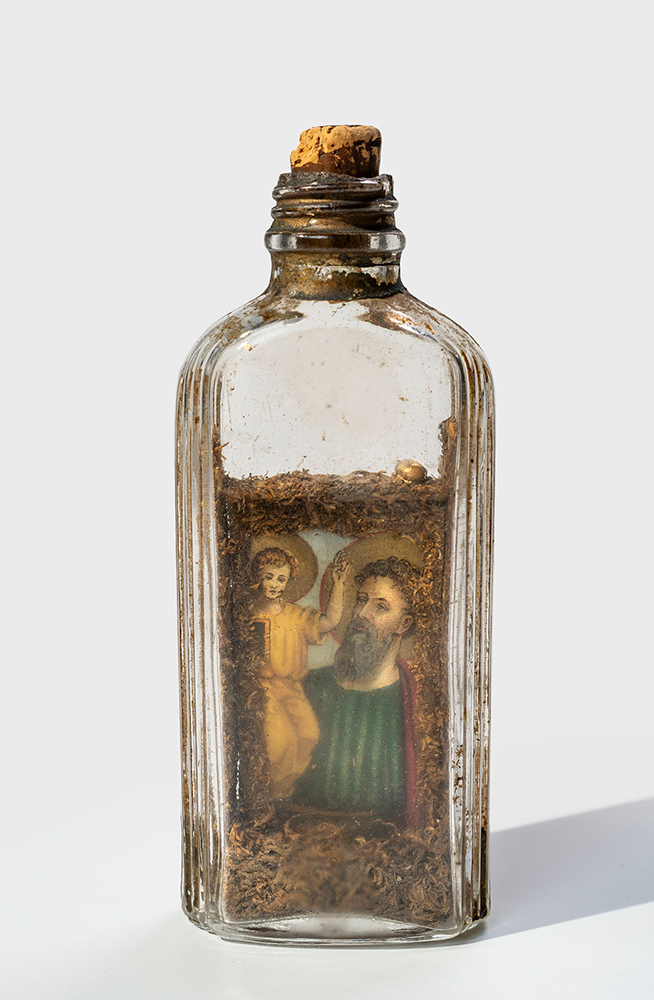 Modern photograph of a glass bottle with cork stopper. One side displays the image of St. Joseph and Christ Child. Images are decorated with dried tobacco on all sides. A small golden bead sits on top of the icons and it appears others used to be present.