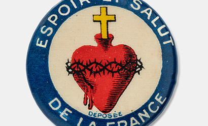Modern photograph of a circular pin depicting a human heart topped with a cross and surrounded by thorns. The blue border has white writing on it.